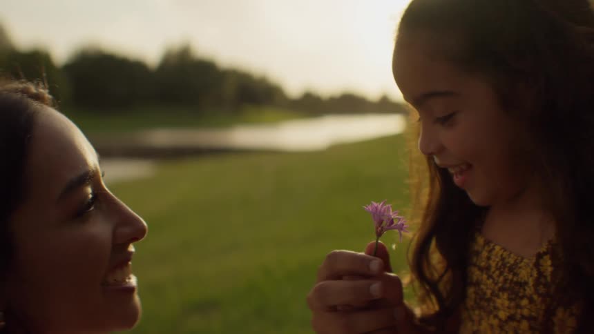 A loving mother watches her young daughter smell a small purple flower during a beautiful sunset in the park