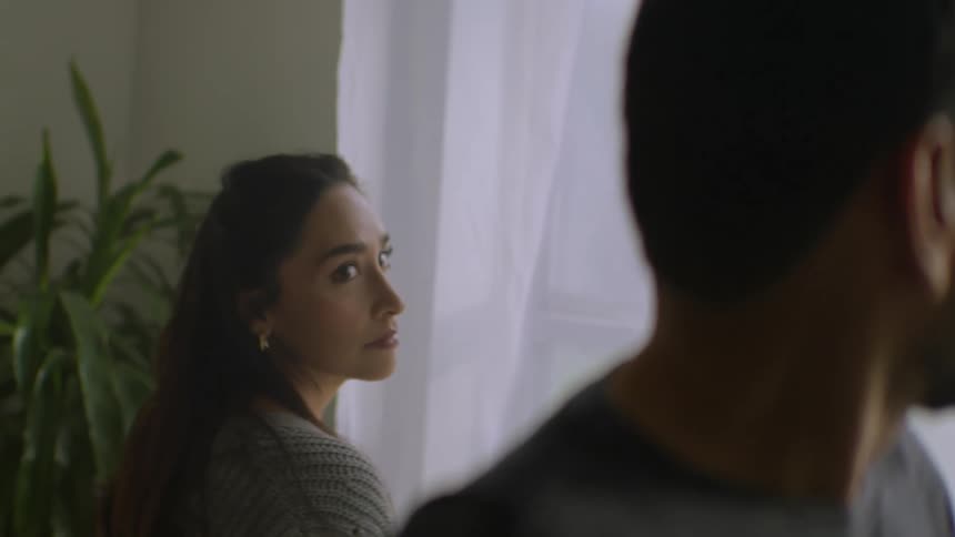 A woman with a concerned expression gazes at her distant partner who stands with his back turned to her