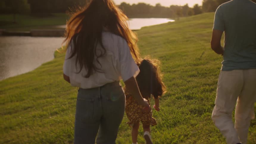 A family with a young daughter is filmed from behind in slow motion as they run through a grassy park next to a lake at golden hour