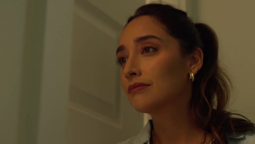 A sad young woman with a melancholic expression leans her head against a door frame in a dimly lit room