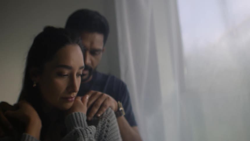 A supportive man gently consoles a pensive woman as they stand together in the soft light of a window
