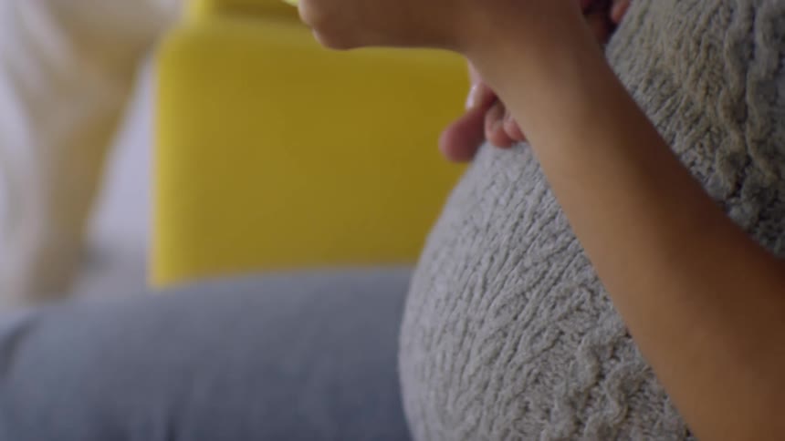 A happy pregnant woman smiles as she holds a mug and her partner lovingly places their hand over hers on the baby bump