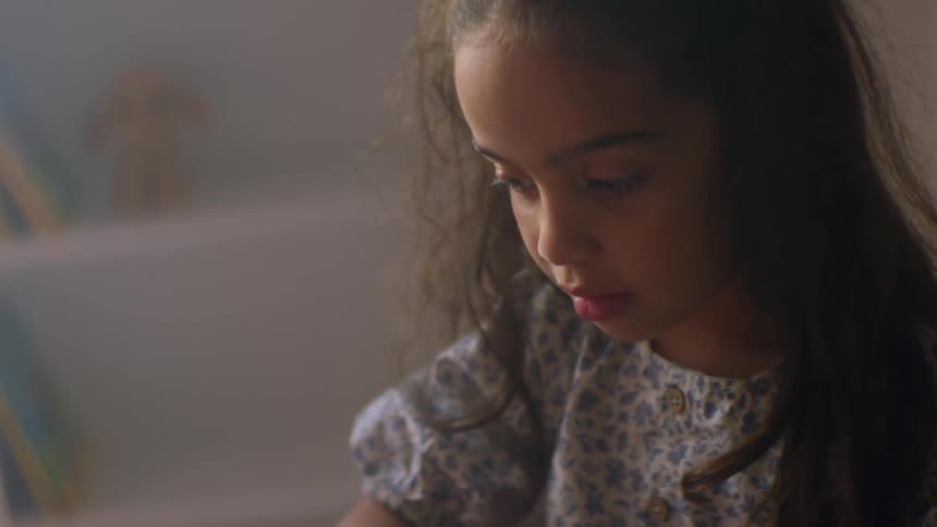 A close-up shot of a young girl with curly hair softly lit as she plays with her pink toy piano on her bed