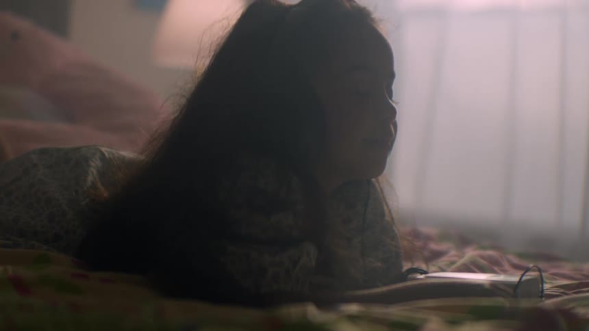 A young girl with long hair lies on her bed in a softly lit room, peacefully listening to music on a portable cassette player with headphones