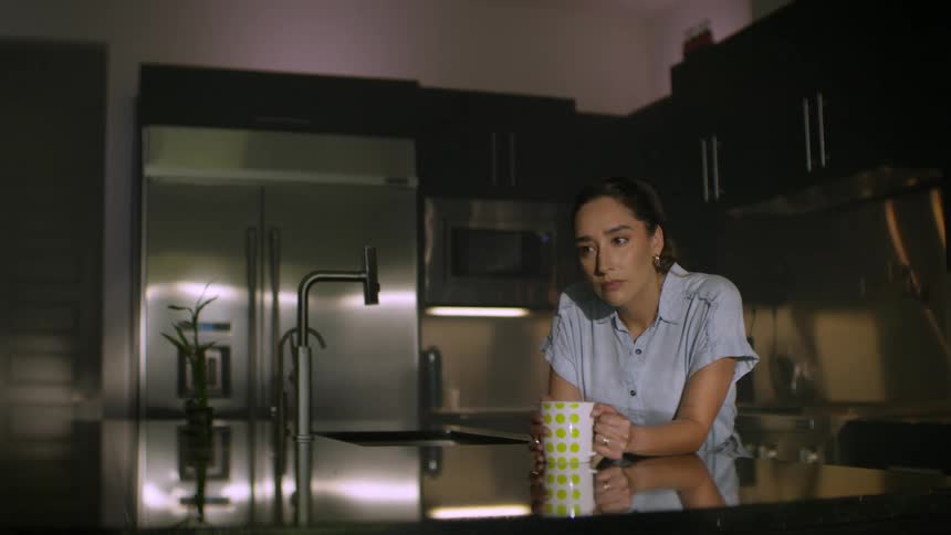 A lonely and pensive woman sits with a mug in a dark, modern kitchen, looking sad and lost in thought