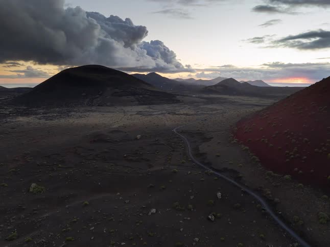 An aerial view pulls back over a vast and desolate volcanic landscape, revealing a red mountain and a winding road under a dramatic, cloudy sunset