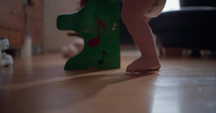 A baby holds onto a toy piano to take their first wobbly steps on a hardwood floor
