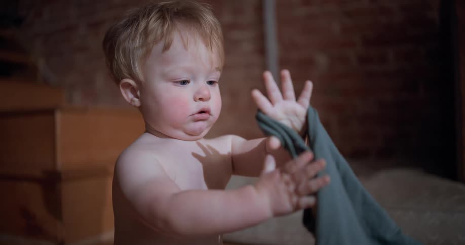A curious blonde toddler with rosy cheeks sits on the floor and intently examines a green hooded sweatshirt in slow motion
