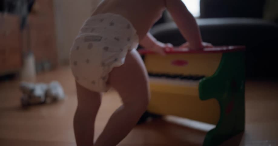 A baby in a diaper stands at a toy piano on the wooden floor of a sunlit living room