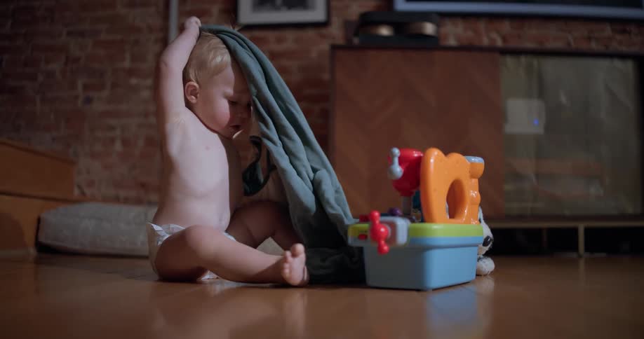 A cute baby boy in a diaper sits on a cozy hardwood floor, playing with a soft blanket as he gets sleepy