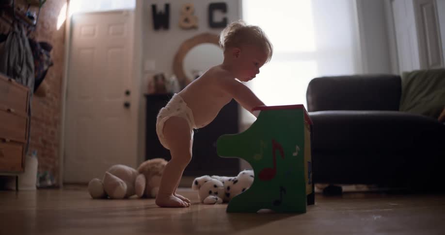 A determined toddler wearing only a diaper learns to walk by pushing a colorful toy piano across the living room floor