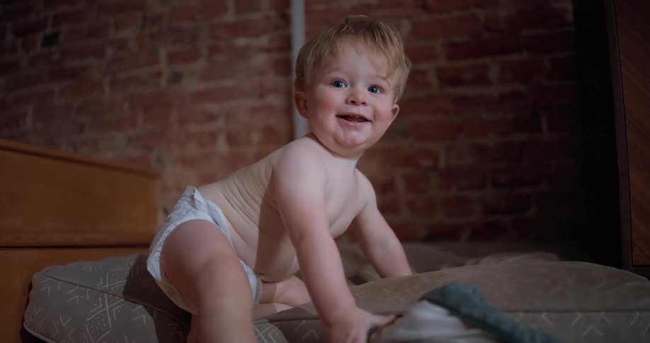 A happy toddler in a diaper plays on a floor cushion and smiles at the camera in slow motion