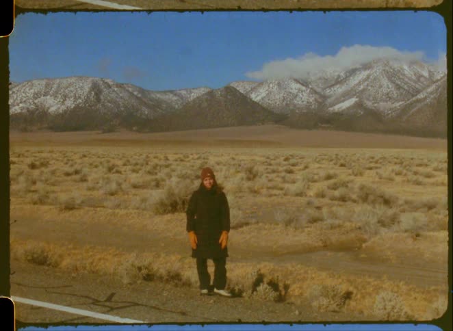 A woman stands in a desert valley before snow-capped mountains in this vintage home movie clip