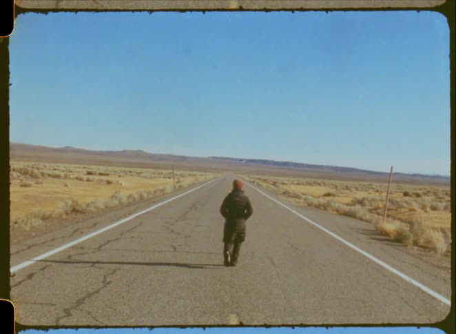 Vintage Super 8 footage shows a woman in a winter coat walking down the middle of a long, empty road in a vast, desolate landscape