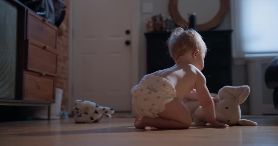 A sweet baby wearing a diaper sits on a sunlit hardwood floor and plays with a stuffed animal in slow motion