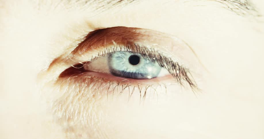 An extreme close-up shot of a person's blue eye opening slowly as a single, dark tear begins to fall