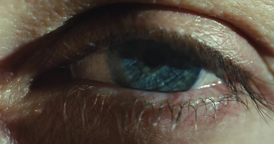 An extreme macro shot captures a blue eye slowly opening, revealing its intricate iris and pupil in dramatic lighting