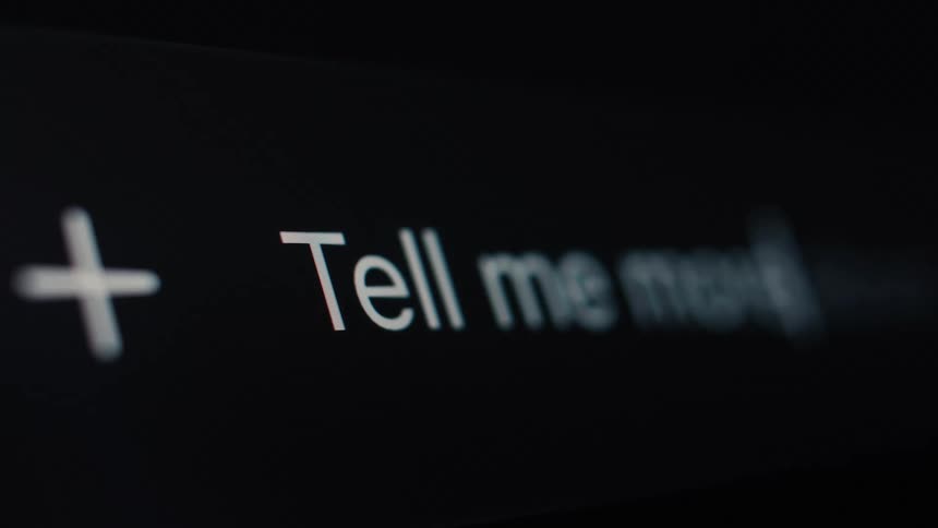 A macro shot shows a user typing the phrase 'Tell me more' into an AI chat prompt on a computer screen