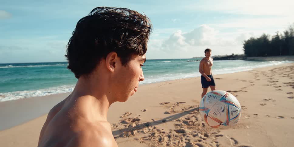 A soccer ball is suspended in mid-air in super slow motion as two young men play on a sunny tropical beach