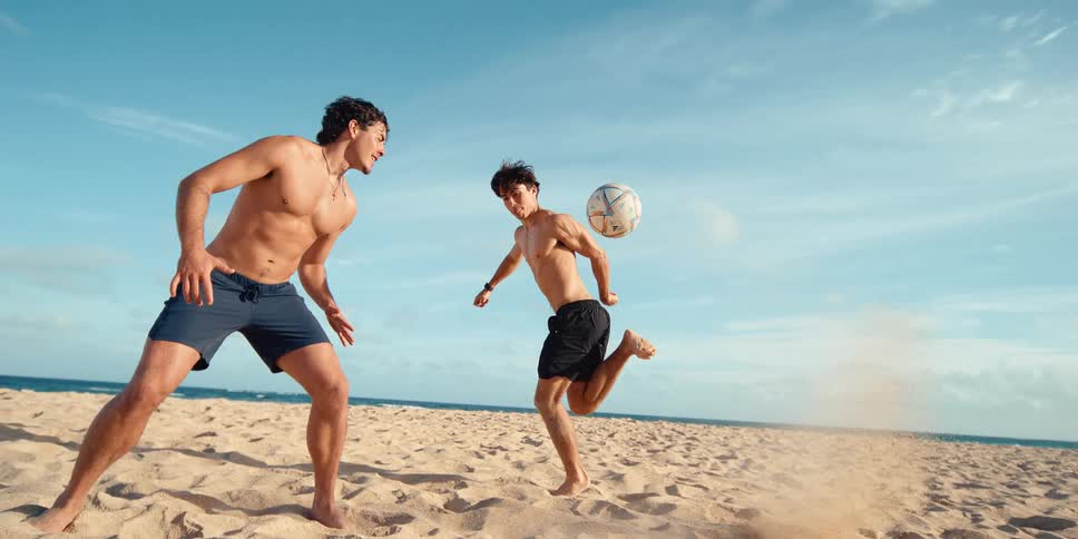 A young man kicks a soccer ball up from the sand while playing with a friend on the beach in super slow motion