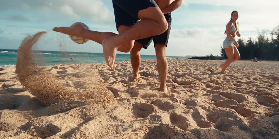 Close-up slow motion shot of a man's bare foot kicking a soccer ball on the beach, sending sand flying as friends play in the background