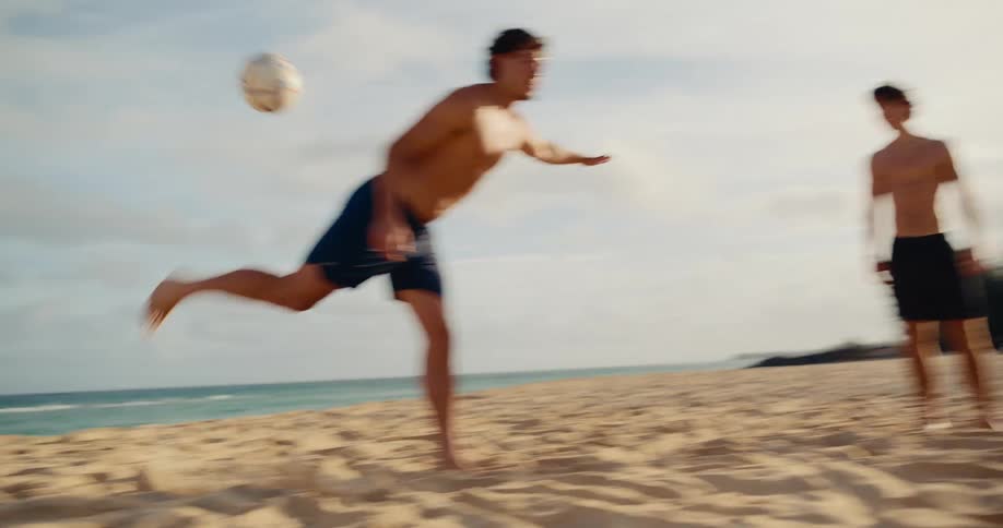 In slow motion, a group of athletic friends kick a soccer ball back and forth on a sandy beach