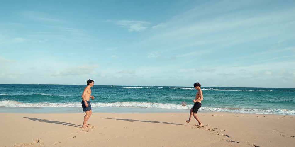 Two athletic young men in swim trunks play with a soccer ball on a sandy beach next to the beautiful turquoise ocean