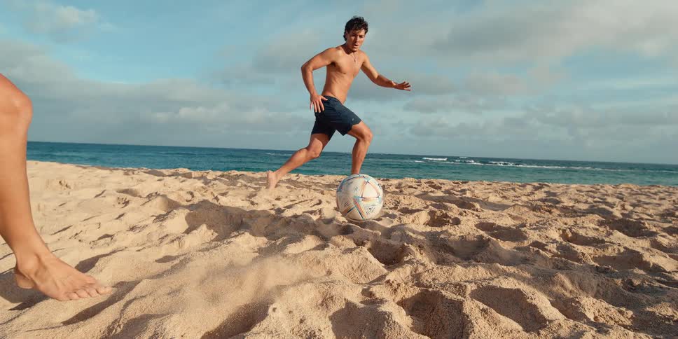 In a super slow motion shot, a woman's foot kicks a soccer ball up from the sand while playing with a man on a tropical beach