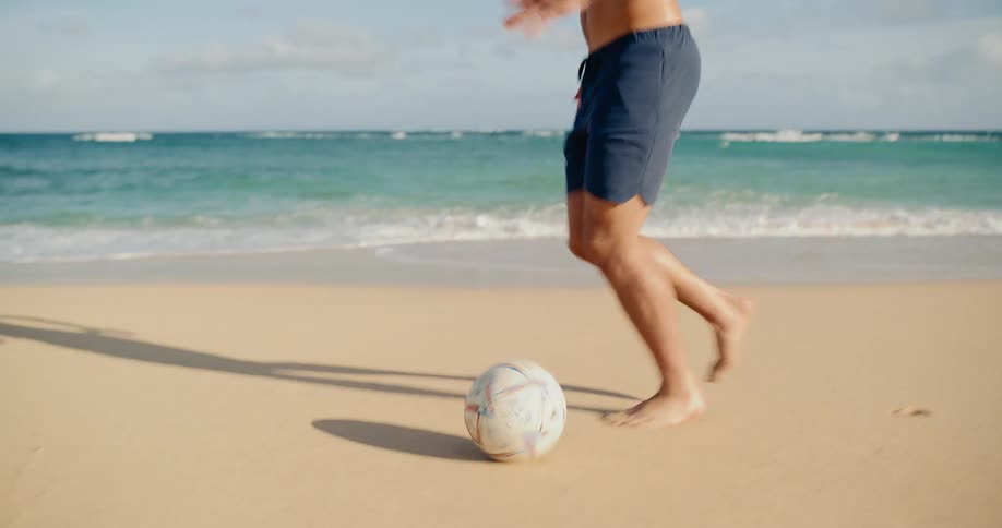 Young athletic man enjoys a game of soccer on the beach next to the ocean waves in slow motion