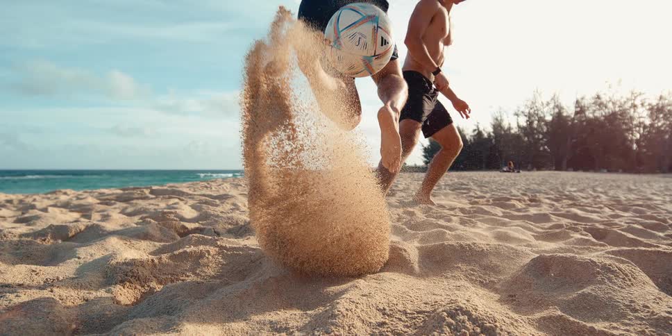 Super slow-motion close-up of a man kicking a soccer ball on a sandy beach, sending a plume of sand flying towards the camera