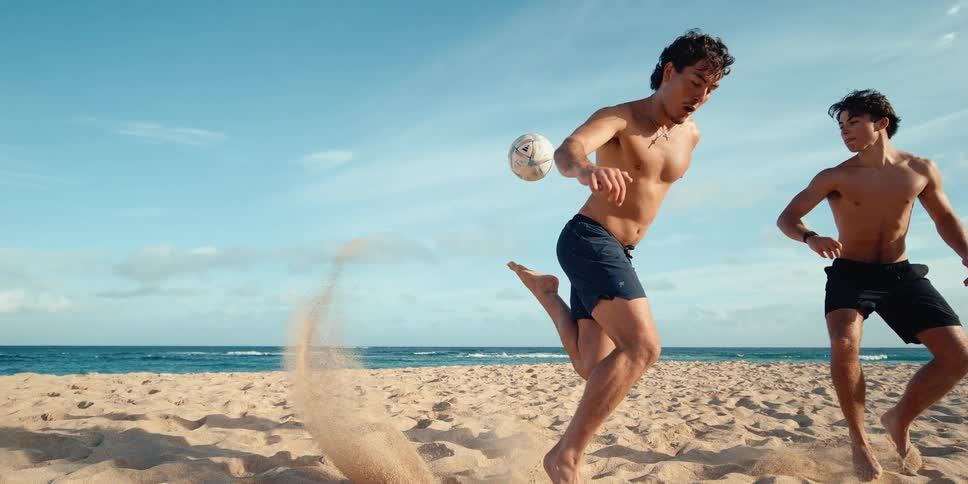 A young man powerfully kicks a soccer ball on a sandy beach, captured in dramatic ultra slow motion as sand explodes from his foot