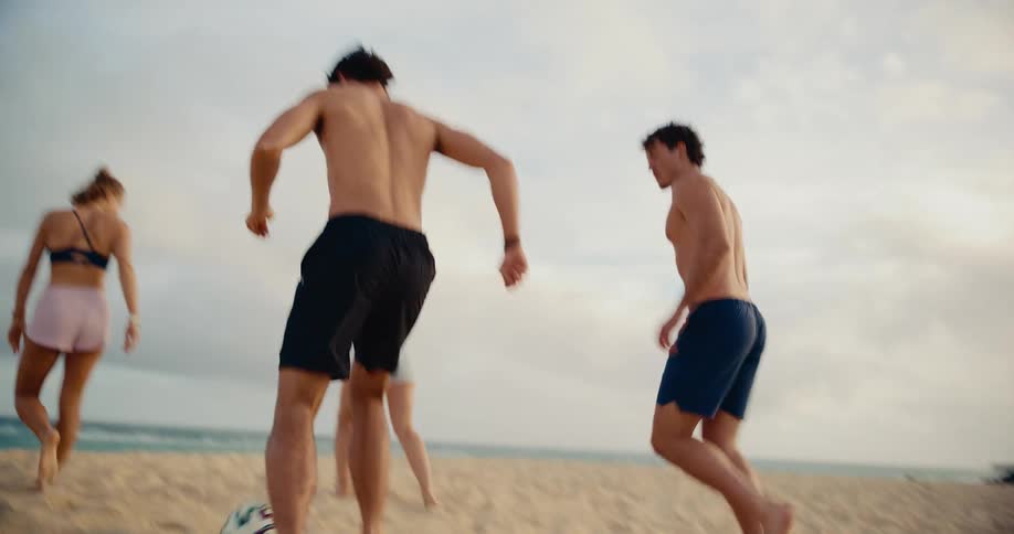 Slow motion shot of active young friends playing soccer barefoot on the sand by the ocean