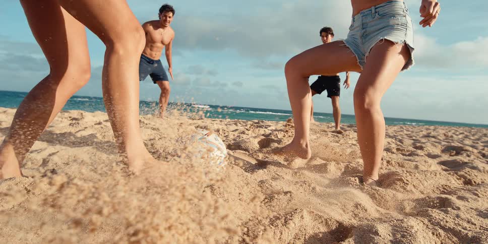 Exciting slow-motion shot shows friends playing soccer on the beach as a woman kicks the ball, sending sand flying towards the camera
