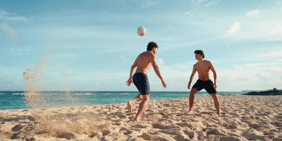 An athletic man kicks a soccer ball on the beach sending sand flying in a dramatic super slow motion shot
