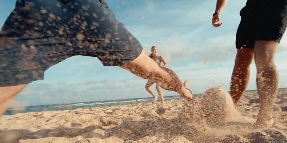 A man kicks a soccer ball on a sandy beach in an extreme slow-motion shot, sending sand flying towards the camera