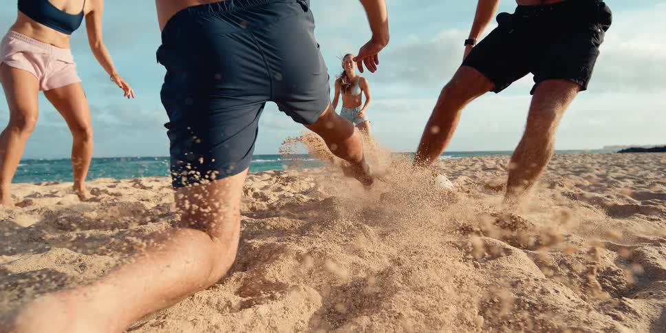 Friends play an energetic game of soccer on a sandy beach with the ocean in the background in dramatic slow motion