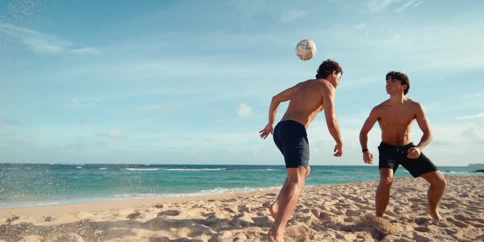 Super slow motion shot of a man kicking a soccer ball on a sandy beach next to the ocean while his friend watches