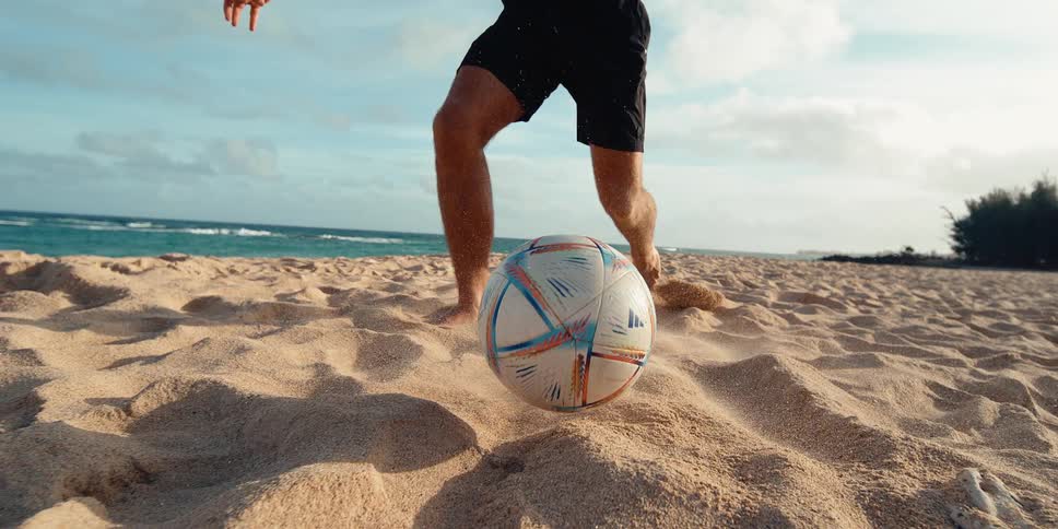 Low angle, super slow motion shot of a man kicking a soccer ball on a sandy beach next to the ocean