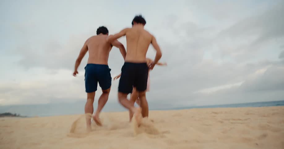 A group of active young friends laugh and play a competitive game of soccer in the sand on the beach in slow motion