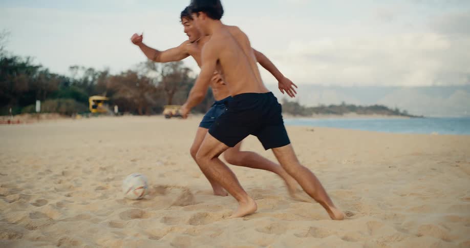 Two athletic young men enjoy a competitive game of soccer barefoot in the sand on a tropical beach in slow motion