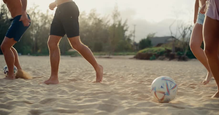 A group of friends' legs are shown in slow motion as they run and dribble soccer balls on a sandy beach at sunset