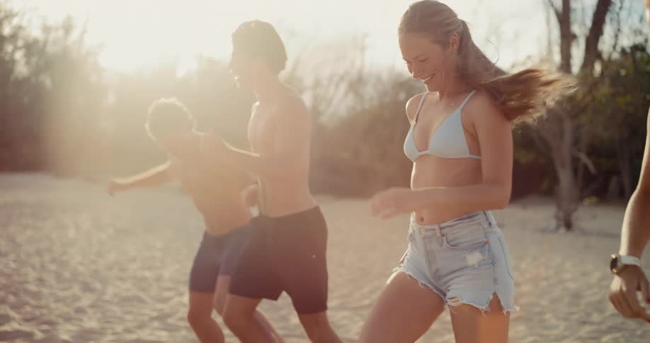 Happy friends laugh and play soccer together on a sandy beach at sunset in slow motion