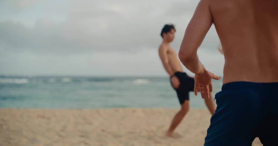 Two athletic young men in swim trunks skillfully play soccer on a sandy beach in slow motion, juggling the ball by the ocean