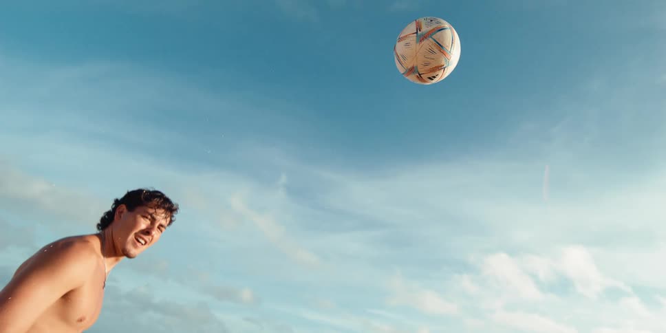 A young man on a sunny beach kicks a soccer ball towards the sky in an epic super slow motion shot