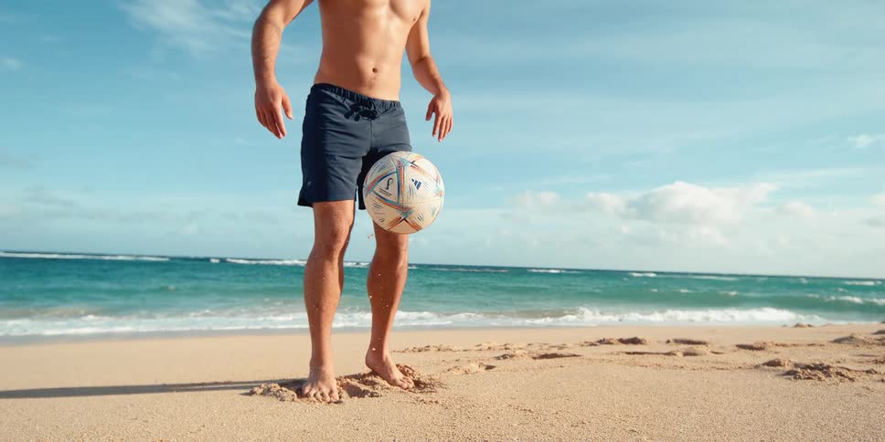 A shirtless, athletic man skillfully balances a soccer ball on his foot in a mesmerizing slow-motion cinemagraph on a sandy beach
