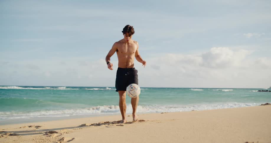 Shirtless athletic man skillfully juggles a soccer ball in slow motion on a sandy beach by the ocean