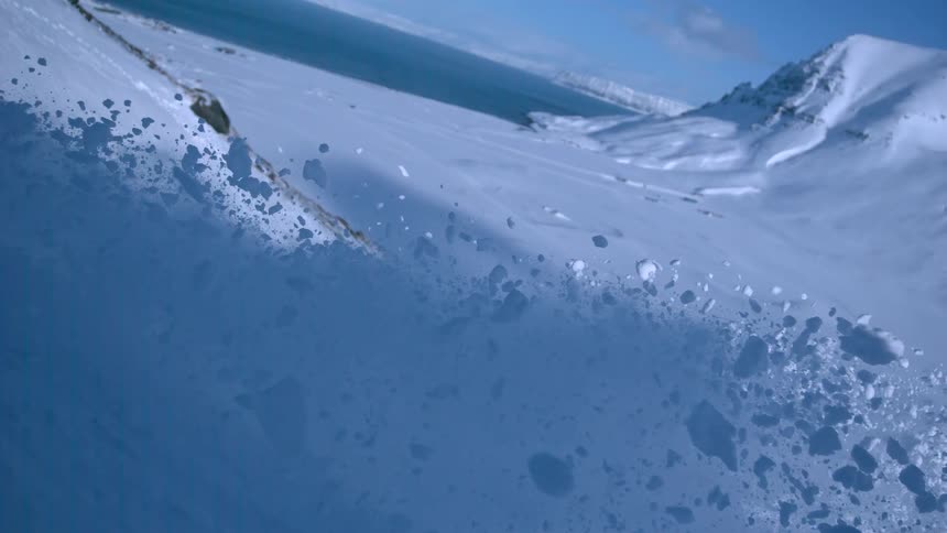 Snow tumbles down a vast mountain slope in slow motion overlooking an icy fjord