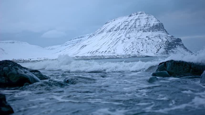 An icy ocean wave crashes in super slow motion towards the stunning snow-covered Kirkjufell mountain in a dramatic Icelandic winter scene