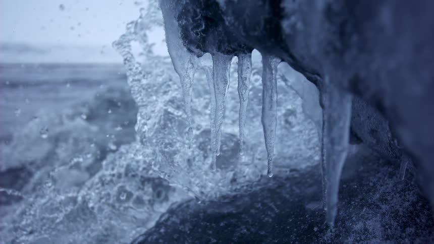 Dramatic slow motion close-up of waves crashing against an icy shore, with icicles hanging in the foreground