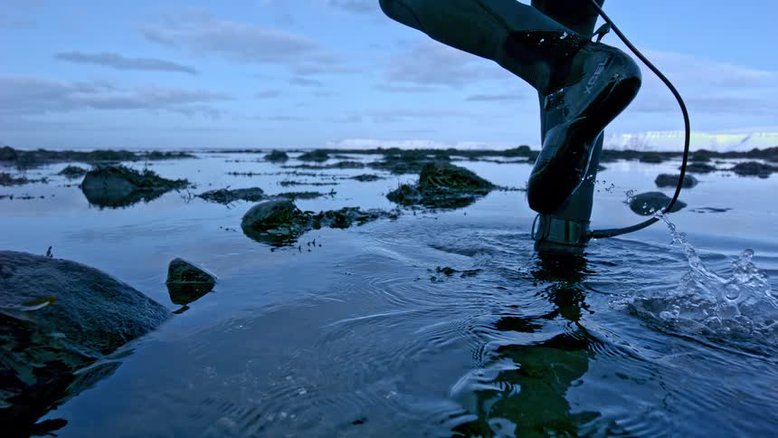 A surfer wearing a wetsuit and booties stands still in the shallow, cold water of a rocky shoreline in dramatic slow motion